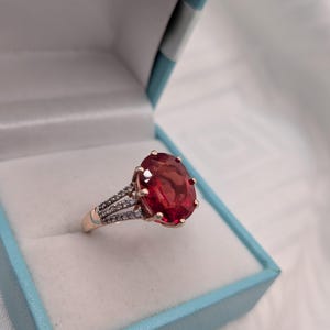 Peut inclure: Une bague ornée d'une grande pierre précieuse rouge foncé de taille ovale, maintenue par six griffes. L'anneau est fabriqué dans un matériau métallique et comporte trois rangées de petites pierres claires et scintillantes sur les côtés.