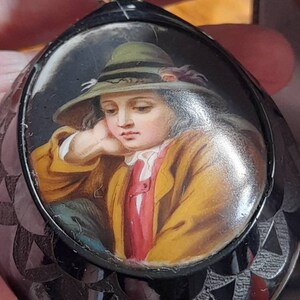 May include: A black and silver locket with a painted portrait of a young person wearing a green hat. The portrait is set in a round, oval-shaped frame.