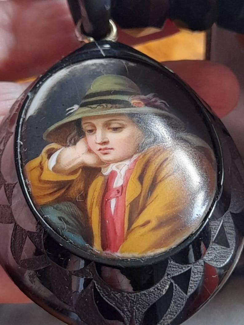 May include: A black and silver locket with a painted portrait of a young person wearing a green hat. The portrait is set in a round, oval-shaped frame.