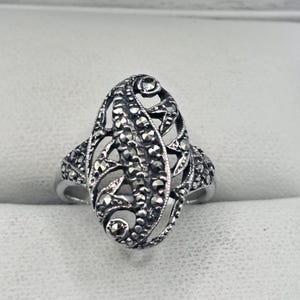 May include: A silver-toned ring with an oval filigree design. The ring features intricate detailing with small, dark stones set within the openwork pattern. The ring's band is smooth and simple, contrasting with the ornate centerpiece.