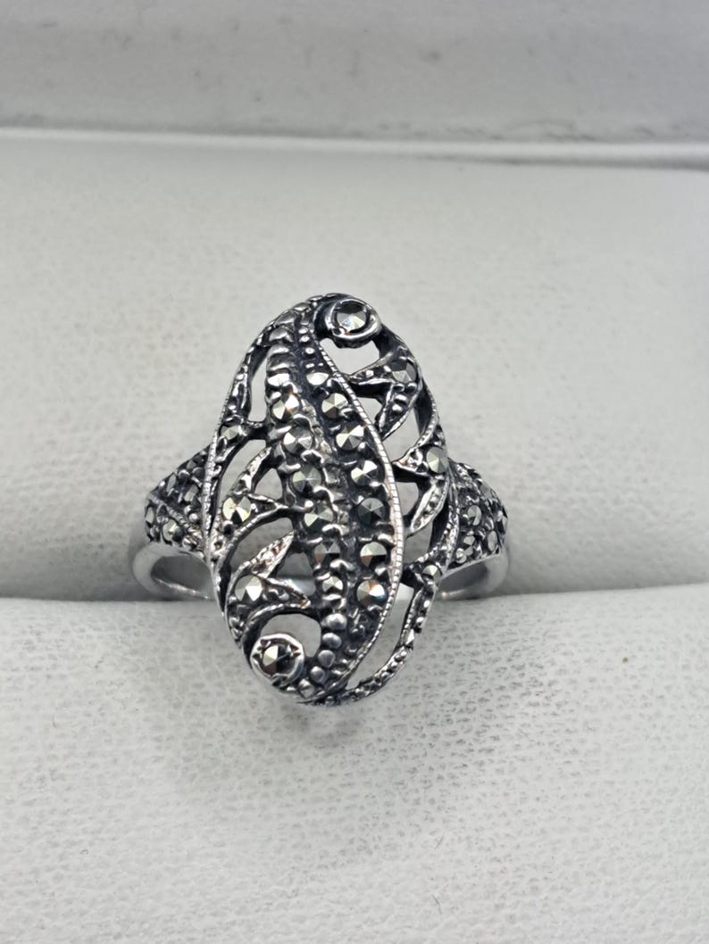 May include: A silver-toned ring with an oval filigree design. The ring features intricate detailing with small, dark stones set within the openwork pattern. The ring's band is smooth and simple, contrasting with the ornate centerpiece.