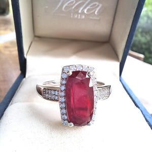May include: A red gemstone ring with a white gold band and a halo of smaller diamonds. The ring is in a blue velvet box with the word "Jader" and the year "1919" printed on the inside.