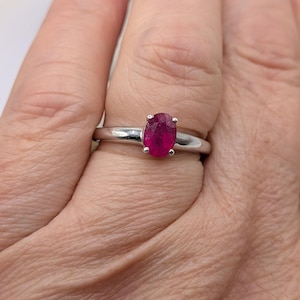 May include: A silver ring featuring a vibrant, oval-cut ruby gemstone. The ruby is a deep, rich red color, set in a simple, polished silver band. The ring is shown on a person's finger.