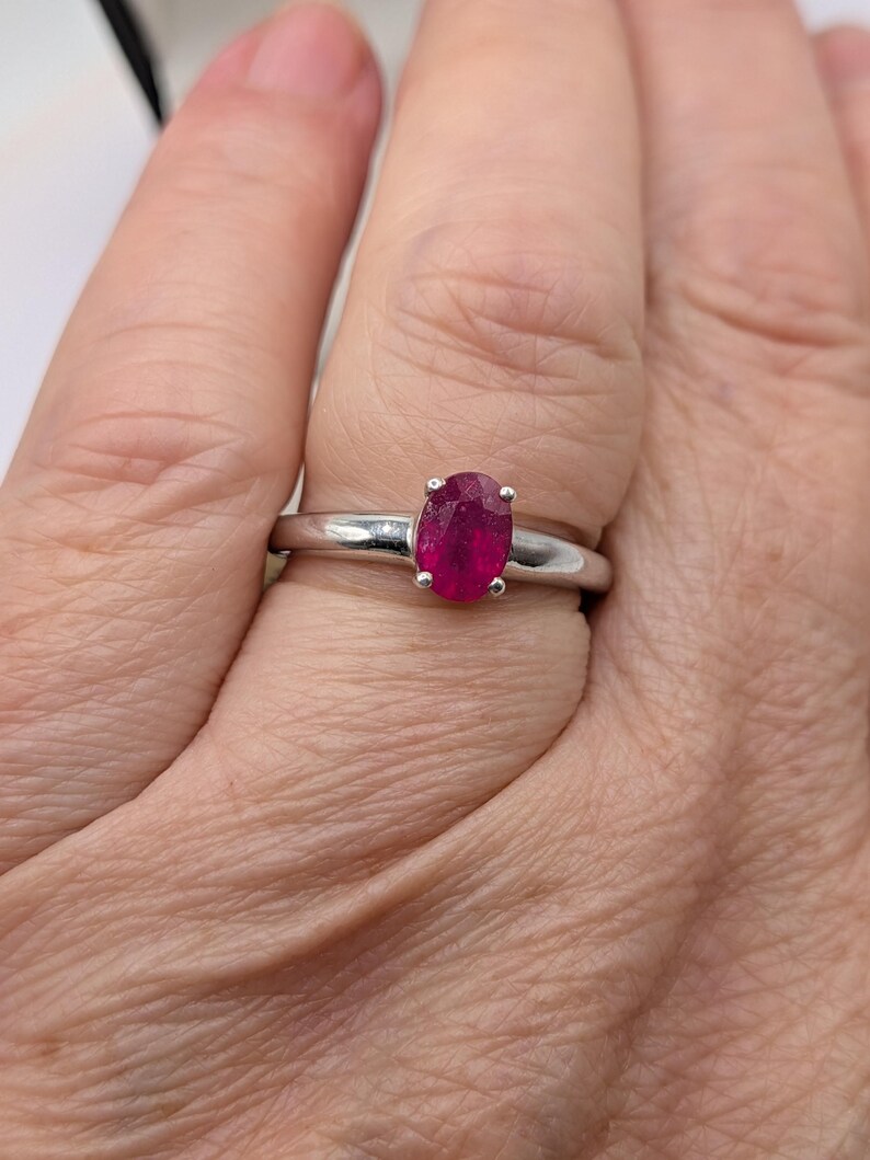 May include: A silver ring featuring a vibrant, oval-cut ruby gemstone. The ruby is a deep, rich red color, set in a simple, polished silver band. The ring is shown on a person's finger.