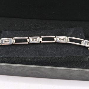Puede incluir: Una pulsera de eslabones en tono plateado que presenta una serie de marcos metálicos rectangulares, cada uno con una pequeña piedra transparente de corte rectangular engastada en el centro. La pulsera alterna eslabones y piedras.