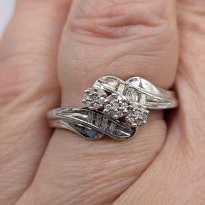 May include: A silver ring with three clusters of small, round diamonds and rectangular diamonds. The ring has a swirling design and is set against a light background.