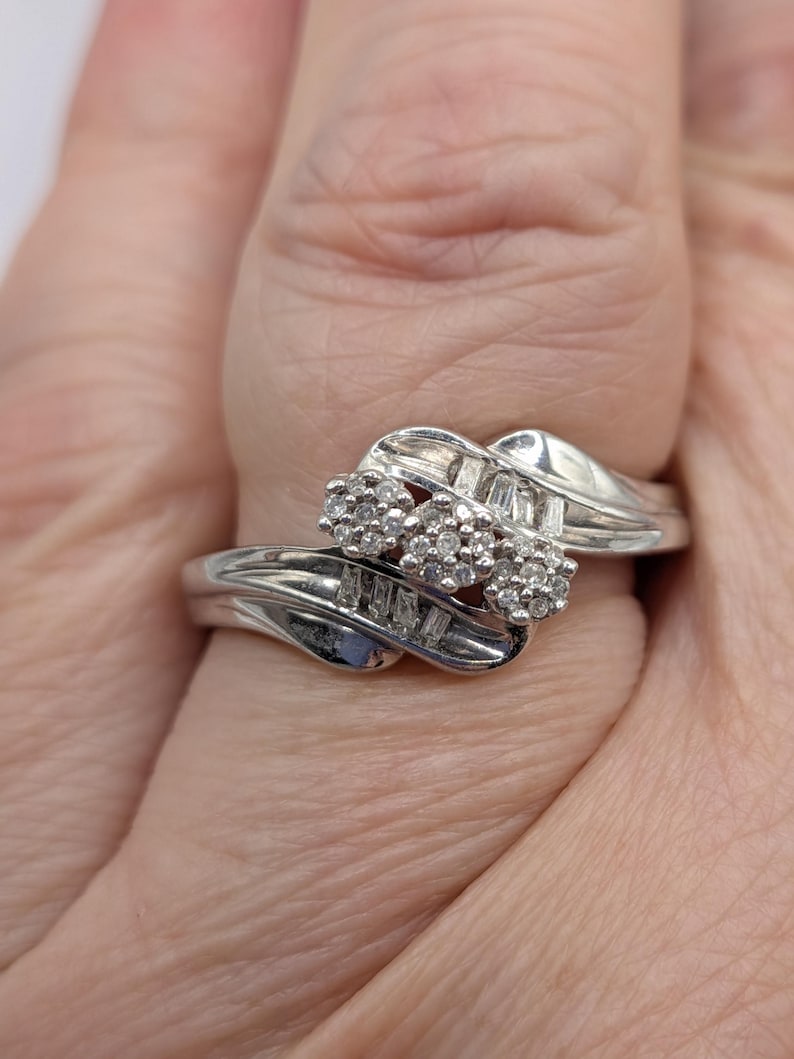 May include: A silver ring with three clusters of small, round diamonds and rectangular diamonds. The ring has a swirling design and is set against a light background.