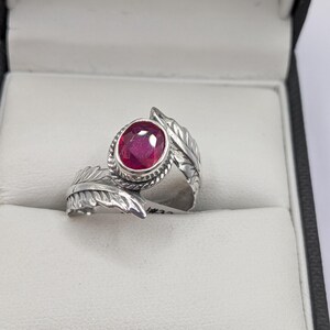 May include: A silver ring with a large, round, ruby-red gemstone. The ring features a leaf design on the band and around the gemstone. The ring is displayed in a black and white jewelry box.