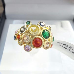 Può includere: Un anello d'oro con un gruppo di pietre preziose colorate. L'anello è incastonato con una varietà di pietre rotonde, tra cui rosse, verdi, blu, gialle e rosa.