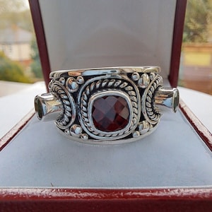 Op de afbeelding: Een zilveren ring met een grote rode edelsteen omringd door kleinere groene edelstenen. De ring heeft een gedetailleerd ontwerp met touwachtige accenten.