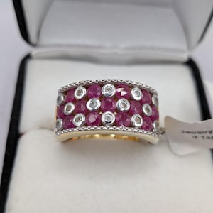 May include: A wide band ring featuring multiple round-cut red rubies and small clear stones set in a silver-toned metal. The stones are arranged in a grid pattern across the surface of the band, which has a textured, beaded edge detail.