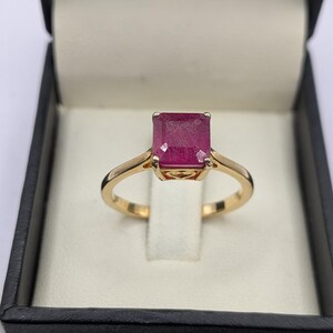 May include: A gold ring featuring a large, rectangular ruby gemstone. The ruby is a deep red hue, set in a gold band. The ring is presented in a black jewellery box with a white interior.