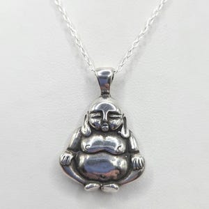 Puede incluir: Un colgante de plata pulida con forma de figura de Buda sentado y sonriente, con los ojos cerrados y el vientre redondeado, suspendido de una delicada cadena de plata. El colgante presenta superficies lisas y reflectantes y rasgos faciales detallados.