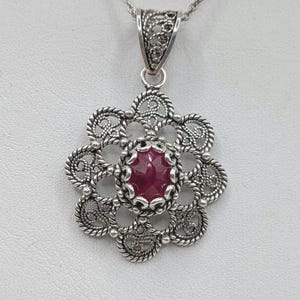May include: A silver pendant necklace with a flower-shaped filigree design. The pendant features a central oval, deep red gemstone. The necklace chain is also silver. The pendant is approximately 3.8 cm in diameter.