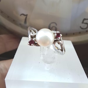May include: A silver ring featuring a large, round white pearl as the centerpiece. Two smaller red gemstones flank the pearl, set within an openwork design. The ring is displayed on a clear, rectangular stand.