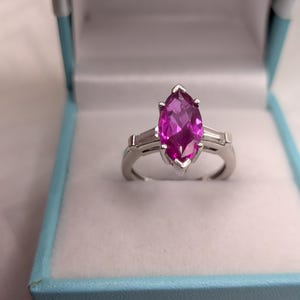 May include: A silver-toned ring featuring a large, marquise-cut vibrant pink gemstone as the centrepiece. The ring is accented by two smaller, rectangular baguette-cut clear stones on either side of the central pink stone, set in a polished metal band.