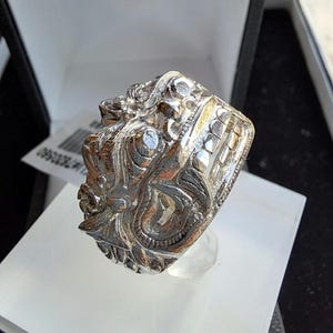 May include: A silver ring featuring an intricate design of a mythical creature's face. The ring is displayed on a white stand.