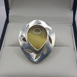May include: A silver ring featuring a pale yellow, teardrop-shaped stone. The ring has a decorative, openwork design with leaf-shaped cutouts. The ring is presented in a dark blue jewellery box.