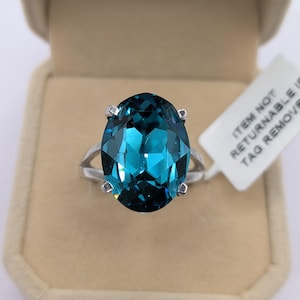 May include: A silver ring featuring a large, oval-cut, teal-colored gemstone. The gemstone is set in a four-prong setting. The ring is displayed in a beige ring box. A white tag with black text is attached to the ring.