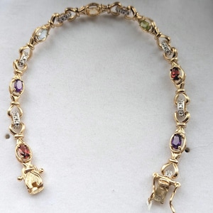 May include: A gold link bracelet featuring alternating oval-cut gemstones in various colours including purple, red, green, and light blue, interspersed with small clear stones set in silver-toned accents, finished with a secure box clasp closure.