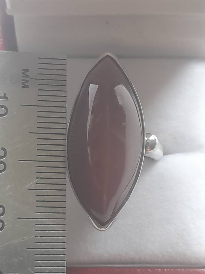 May include: A silver ring with a large, dark red, marquise-shaped gemstone. The ring is next to a ruler, showing the stone is approximately 2.5 inches long.