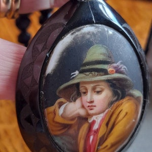 May include: A black cameo pendant with a hand-painted portrait of a young person wearing a green hat. The portrait is set in a round, white cameo.