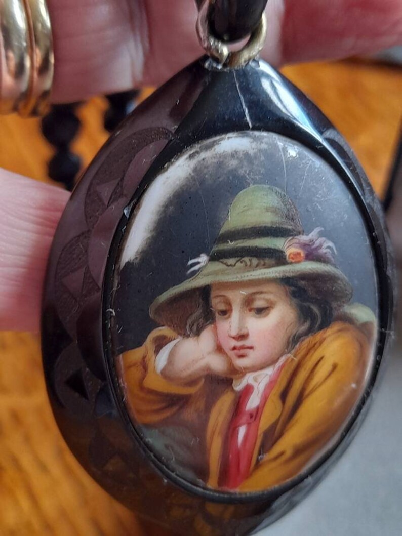 May include: A black cameo pendant with a hand-painted portrait of a young person wearing a green hat. The portrait is set in a round, white cameo.