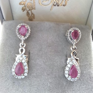 May include: A pair of silver drop earrings with a pear-shaped, deep red ruby center stone, surrounded by a halo of small, clear gemstones. The earrings are displayed in a gray jewelry box.