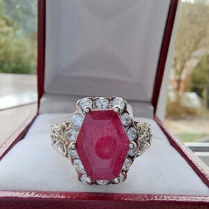 May include: A silver ring featuring a large, hexagonal, red gemstone surrounded by smaller, clear gemstones.