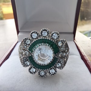 Op de afbeelding: Een zilveren ring met een grote, heldere, ronde edelsteen omringd door kleinere, heldere edelstenen. De ring heeft een groene emailleband rond de middelste steen.