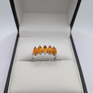 May include: A silver ring set with five marquise-cut, orange-yellow gemstones. The ring is presented in a black jewellery box with a white interior. The gemstones' colour is accentuated by the box's contrasting design.