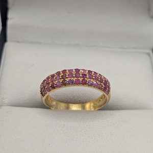 May include: A gold ring featuring three rows of small, round, pink gemstones. The ring has a polished gold band and is presented in a white jewellery box.