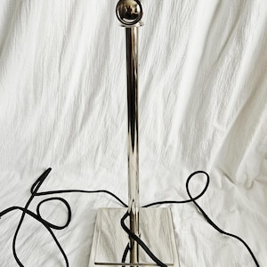 May include: A polished silver floor lamp with a square base and a slender, vertical pole. The lamp features a decorative top with multiple light fixtures. A black electrical cord is visible, adding contrast to the metallic finish.