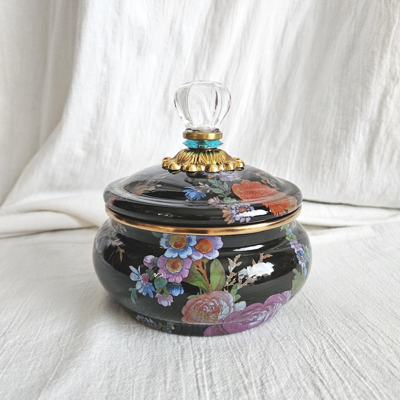 May include: A black ceramic jar with a lid, adorned with a floral pattern in shades of red, purple, blue, and white. The lid is topped with a clear, faceted knob and gold-colored accents. The jar has a gold rim.
