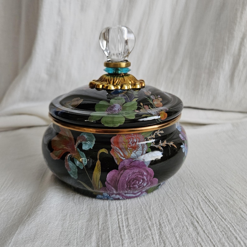May include: A black ceramic jar with a lid, adorned with a floral pattern in shades of purple, pink, and green. The lid is topped with a clear, faceted knob and gold-colored accents. The jar has a gold rim.