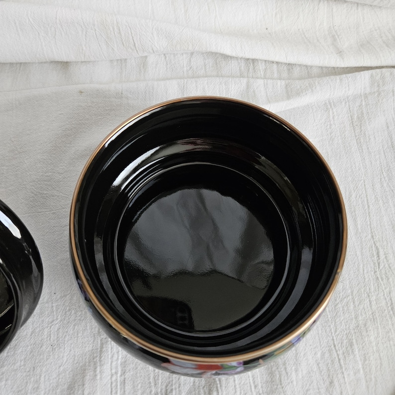 May include: A black ceramic bowl with a glossy finish and a gold rim. The bowl has a wide, open design and a smooth interior. Floral patterns are visible on the exterior.