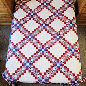 May include: A rectangular quilt featuring a repeating pattern of large white diamond-shaped blocks separated by a grid of smaller squares in red, blue, and multi-colored floral prints. The white blocks have a subtle stitched floral design.