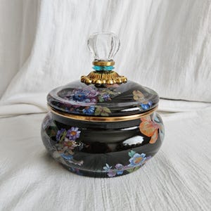 May include: A black ceramic jar with a lid, adorned with a floral pattern in shades of blue, purple, and orange. The lid is topped with a clear, decorative handle and gold accents. The jar has a gold rim.
