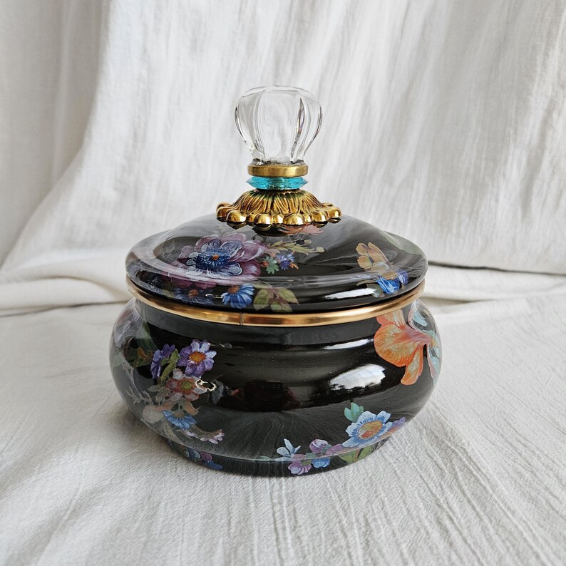 May include: A black ceramic jar with a lid, adorned with a floral pattern in shades of blue, purple, and orange. The lid is topped with a clear, decorative handle and gold accents. The jar has a gold rim.