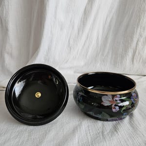 May include: A black ceramic container with a lid, featuring a floral design with white, purple, and blue irises. The rim of the container is accented with gold. The lid has a gold knob.