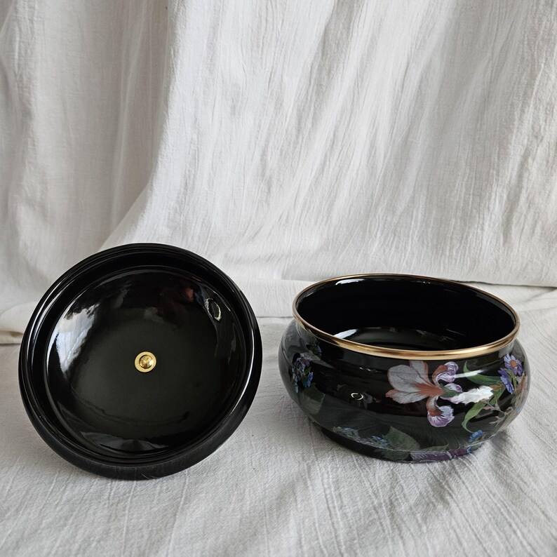 May include: A black ceramic container with a lid, featuring a floral design with white, purple, and blue irises. The rim of the container is accented with gold. The lid has a gold knob.