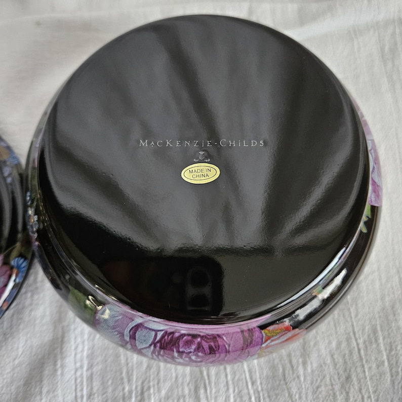 May include: A black, round decorative bowl with a floral design in shades of purple, pink, and white. The bowl has a glossy finish and the text "MacKenzie-Childs" is printed on the top. A small sticker reads "Made in China".
