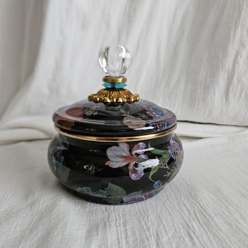 May include: A black ceramic jar with a lid, adorned with floral patterns in shades of white, purple, and green. The lid is topped with a clear, crystal-like knob and gold accents. The jar has a gold rim.