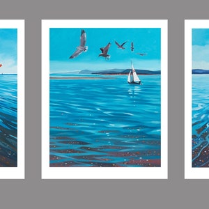 Może przedstawiać: Zestaw trzech obrazów przedstawiających scenę plażową z ludźmi spacerującymi po piasku, żaglówką na wodzie i mewami latającymi nad głowami. Obrazy są oprawione w białe ramki i mają niebieski tło.