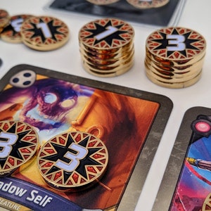 May include: A close-up of a card game with a card titled "Shadow Self" featuring a creature with a skull-like face. The card is surrounded by gold and red tokens with numbers on them. The tokens are stacked in piles of different heights.