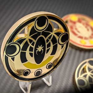 May include: A gold-colored metal pin with a black and yellow design. The design features a stylized spider web with three crescent moons and a spider in the center. The pin is on a clear acrylic stand.