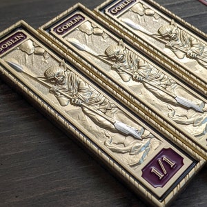 Peut inclure: Trois jetons rectangulaires dorés avec un motif de gobelin et le mot "GOBLIN" en haut. Chaque jeton représente un gobelin tenant une lance et le chiffre "1/1" en bas. Les jetons sont posés sur une surface en bois sombre.