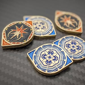 May include: Close-up of several gold-colored, geometric coins with intricate designs. The coins feature a compass rose, starburst, and other geometric shapes in shades of blue, orange, and black. The coins have a shiny, metallic finish and are arranged on a dark surface.