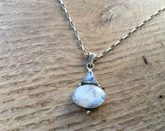 May include: A silver necklace with a pendant featuring two moonstones. The pendant has an oval-shaped moonstone below a triangular one, both displaying a milky white and blue iridescence. The chain is a simple link style.