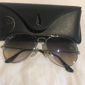 K&ouml;nnte beinhalten: Silberne Aviator-Sonnenbrille mit braunen Verlaufsgl&auml;sern und einem schwarzen Lederetui. Das Etui tr&auml;gt das Ray-Ban-Logo auf der Vorderseite.